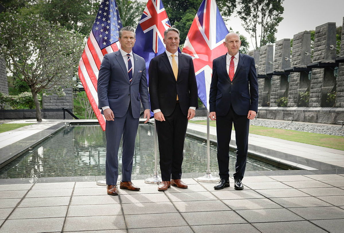 Three suited officials stand by a reflecting pool with US and UK flags in a stone courtyard.