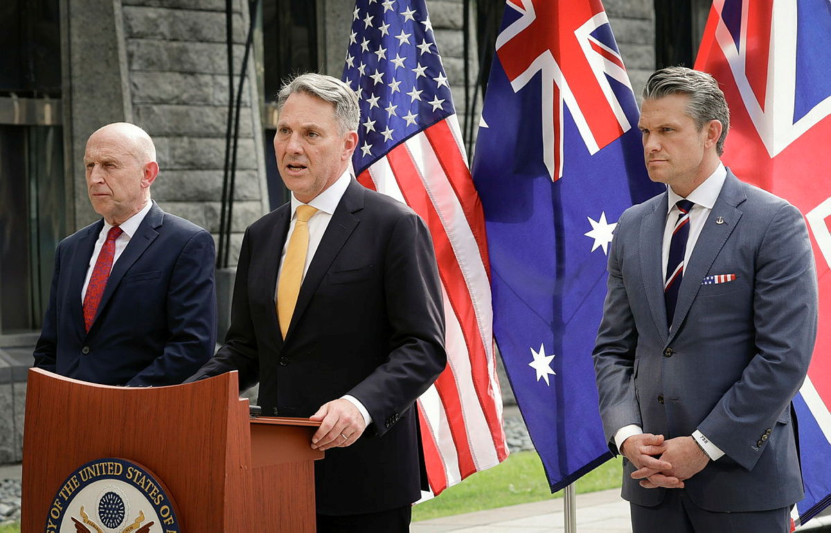 Three officials at a podium speak before US, Australian and UK flags in a stone courtyard.