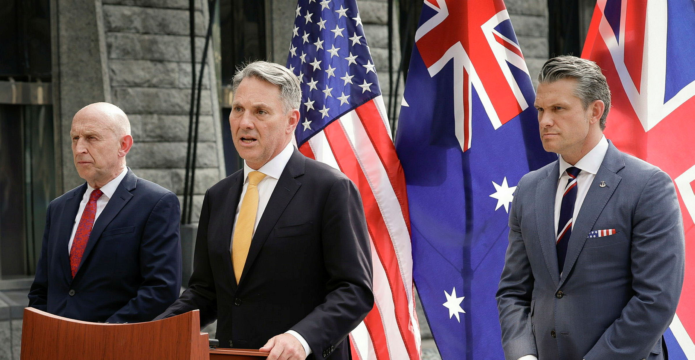 Three officials at a podium speak before US, Australian and UK flags in a stone courtyard.