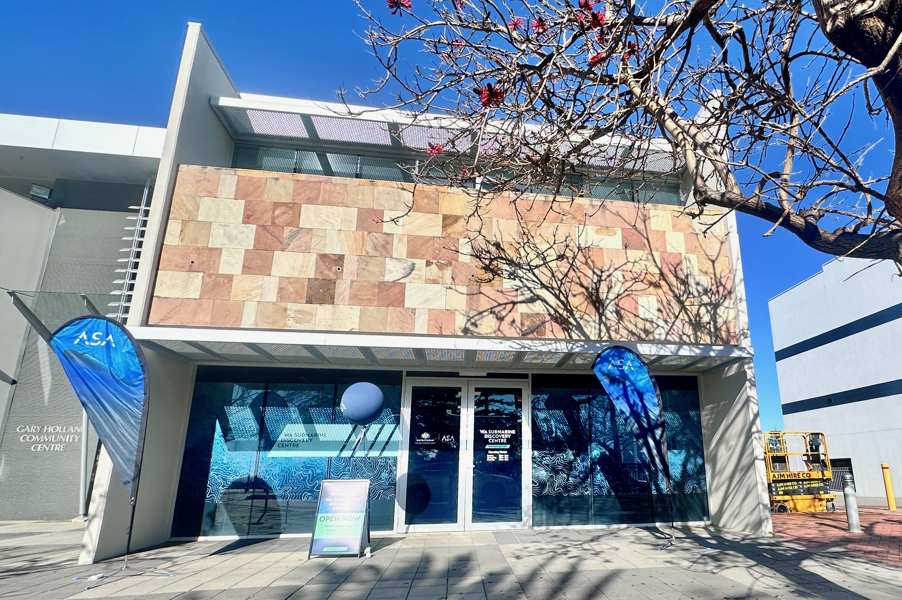 Front entrance of WA Submarine Discovery Centre, with blue window graphics, ASA flags and a tree overhead.