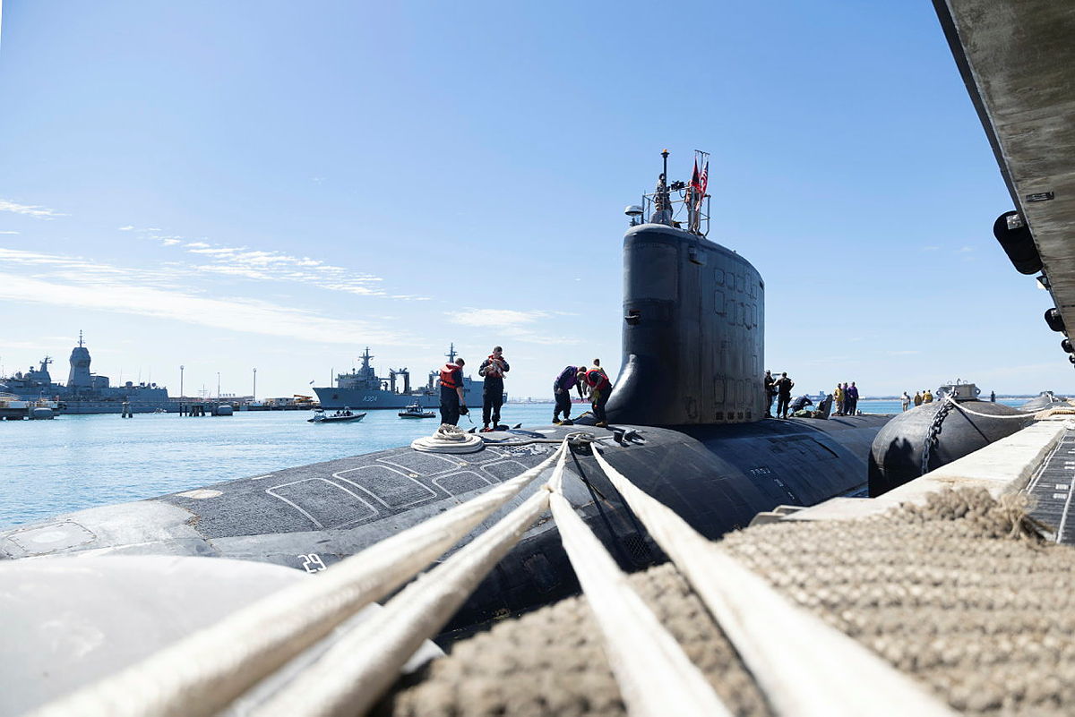 Crew work on a submarine moored at HMAS Stirling, with naval ships and small boats in the harbour.