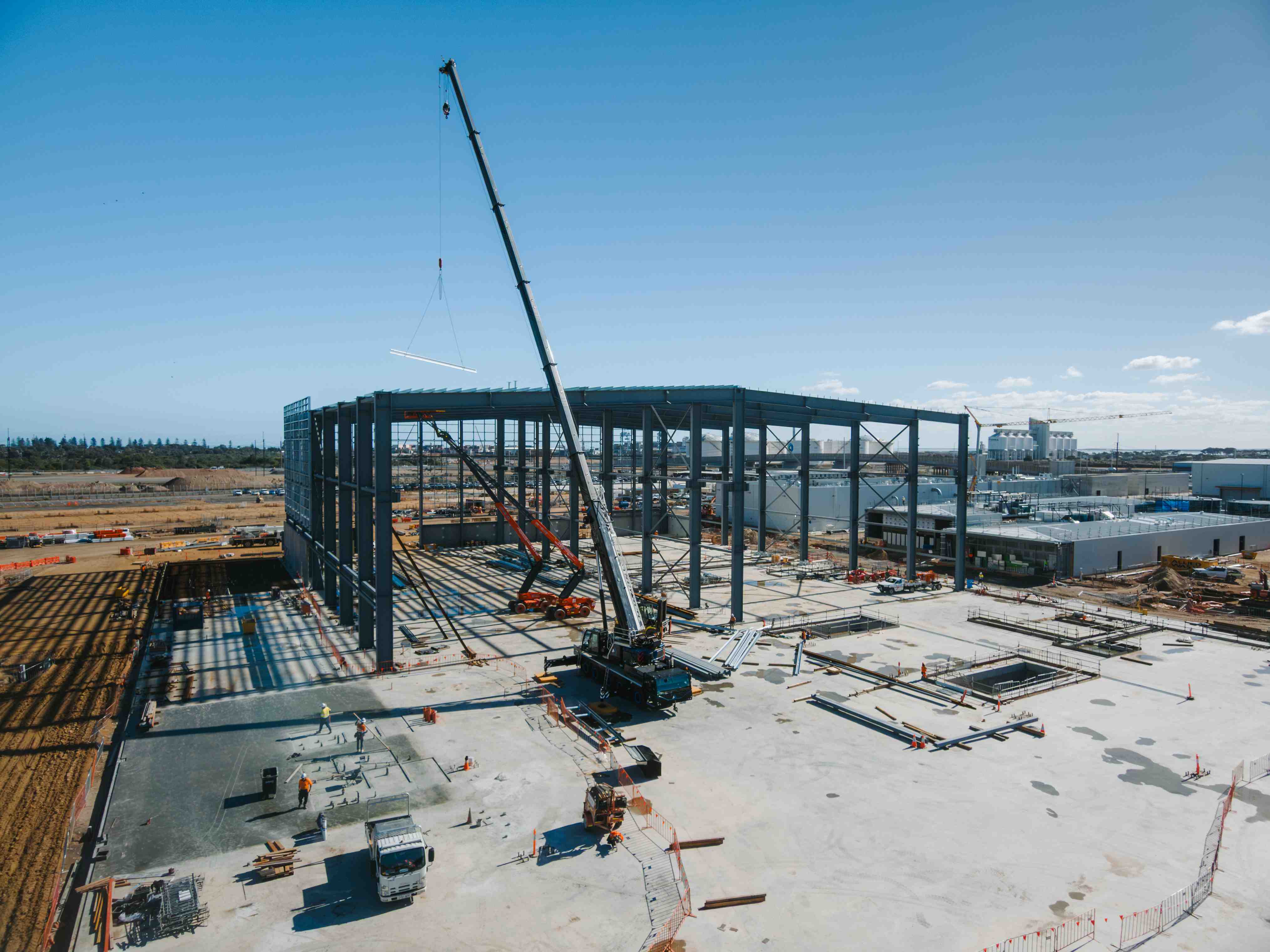 Crane erects a steel frame for a large industrial building at an active construction site.