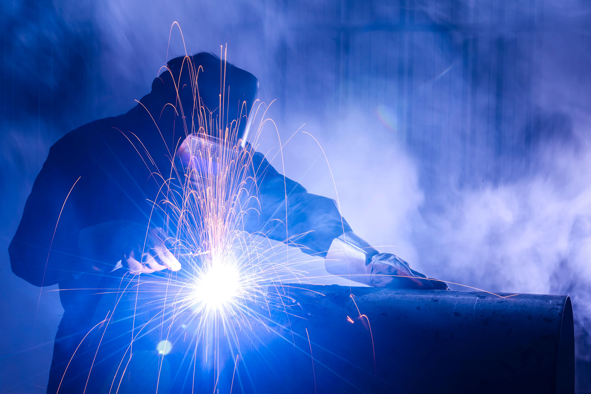 Welder joins a steel pipe, with bright sparks and blue light filling an industrial workshop.