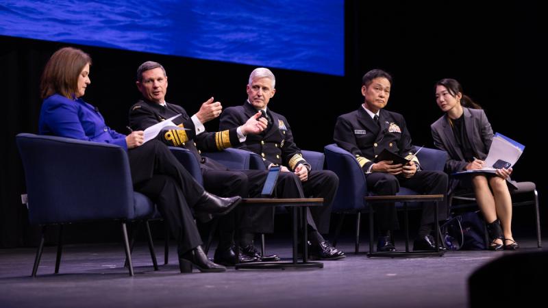 Five speakers take part in a panel discussion on stage, seated before an audience at a conference.