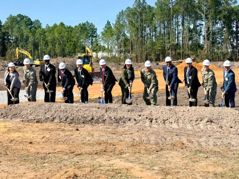 Group of people in hard hats turn the first sod at a construction site, with excavators and trees behind.