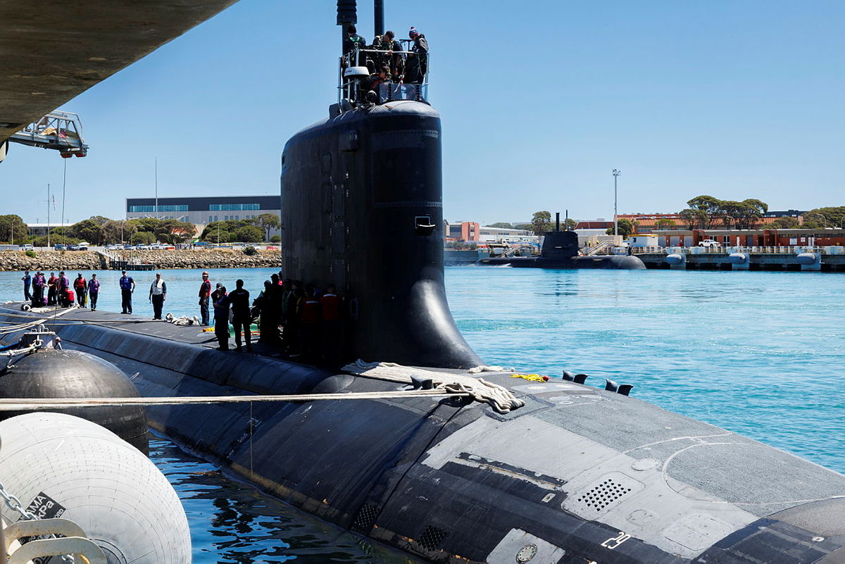 Submarine berthed at a naval base, with crew on deck and another submarine moored across the harbour.