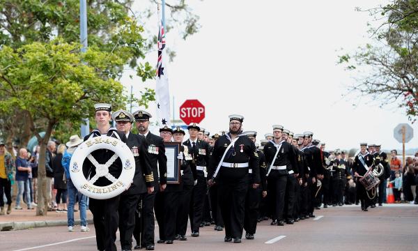 Sailors in black uniforms marching in formation during a parade, led by a member holding an HMAS Stirling life ring