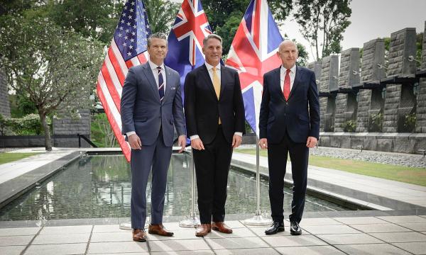 Three suited officials stand by a reflecting pool with US and UK flags in a stone courtyard.