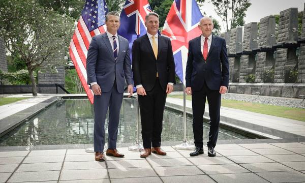 Three suited officials stand by a reflecting pool with US and UK flags in a stone courtyard.