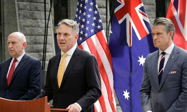 Three officials at a podium speak before US, Australian and UK flags in a stone courtyard.
