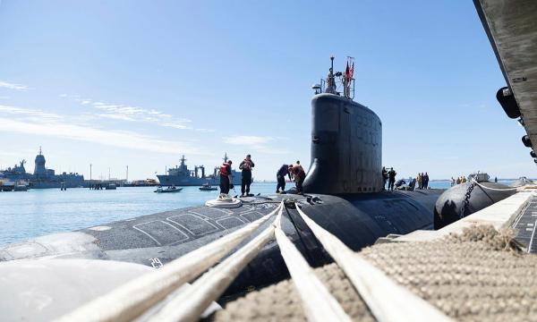 Crew work on a submarine moored at HMAS Stirling, with naval ships and small boats in the harbour.