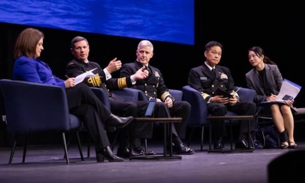 Five speakers take part in a panel discussion on stage, seated before an audience at a conference.