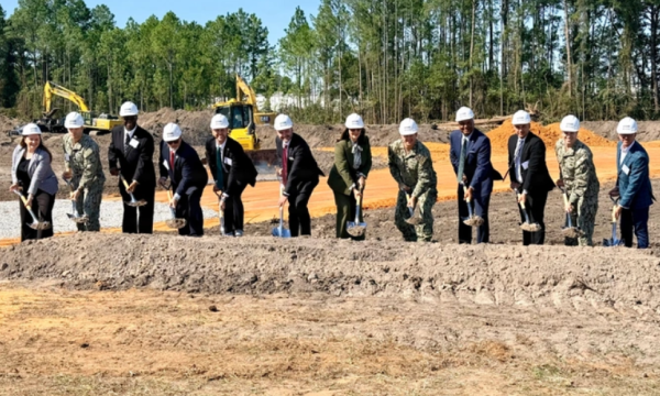 Group of people in hard hats turn the first sod at a construction site, with excavators and trees behind.