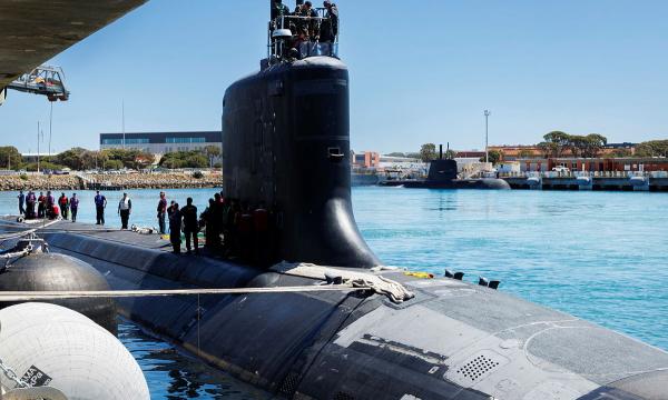 Submarine berthed at a naval base, with crew on deck and another submarine moored across the harbour.