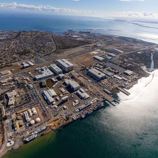 Aerial view of a coastal shipyard with docks, warehouses and parked cars beside a calm bay and residential area.