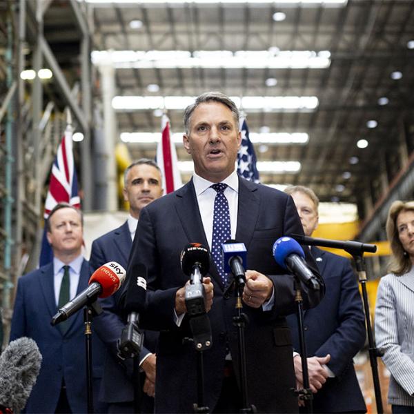 Official speaks at microphones in a large factory, with UK, US and Australian flags and officials standing behind.