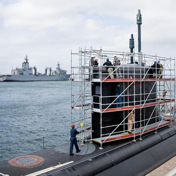 Crew work on a submarine tower under scaffolding at a dock, with naval ships moored in the harbour behind.