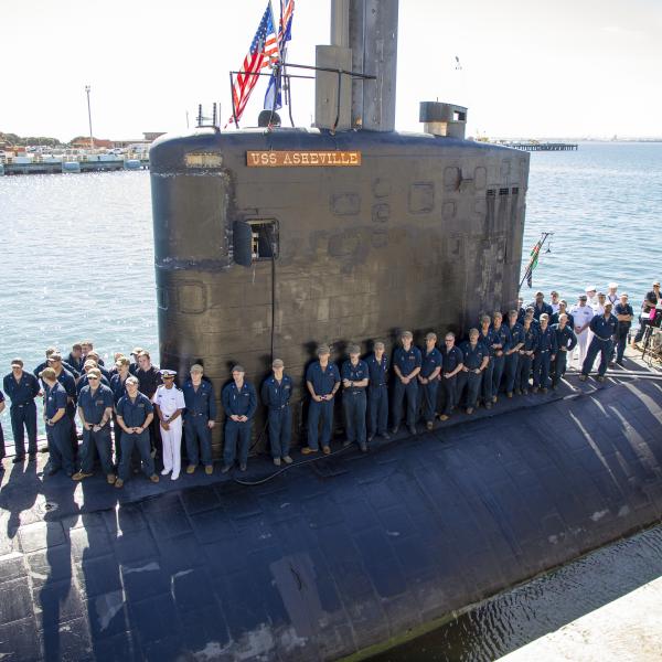 Crew stand in rows on submarine USS Asheville docked at a harbour, with buildings and water in the background.