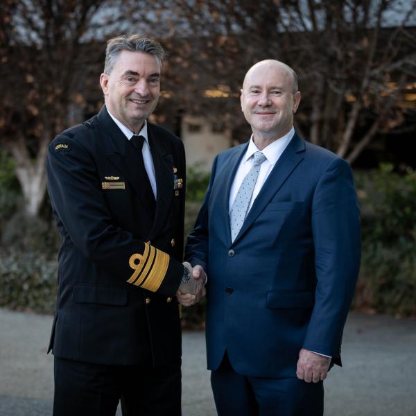 Two people in suit and naval uniform shake hands outdoors, with trees and garden path in the background.