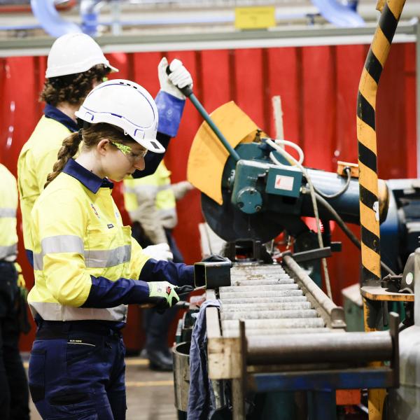 Workers in high vis gear use machinery to cut metal parts on a factory floor with red safety screens behind.
