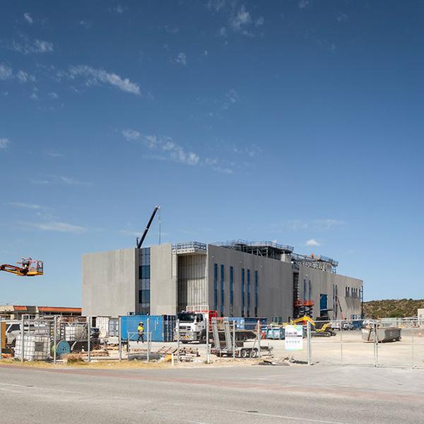 Large industrial building under construction with cranes, trucks and scaffolding behind a fenced work site.
