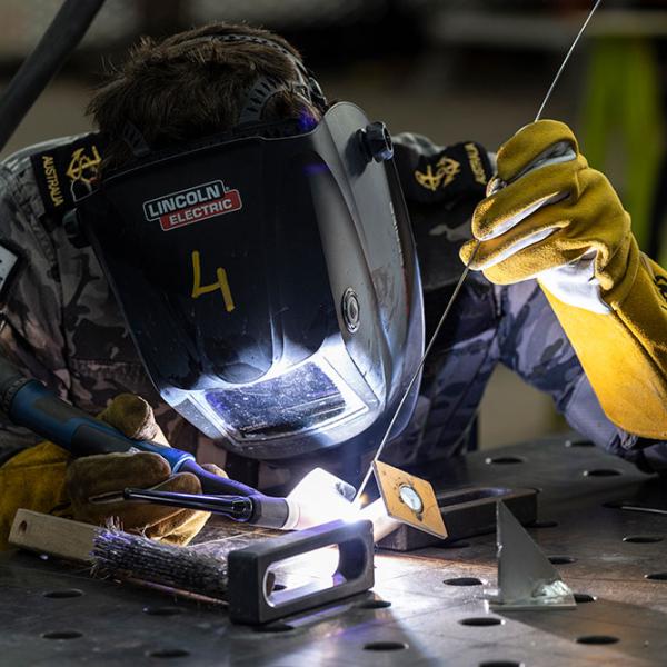 Person welds metal with a torch at a workbench, wearing a protective mask and gloves in a workshop.